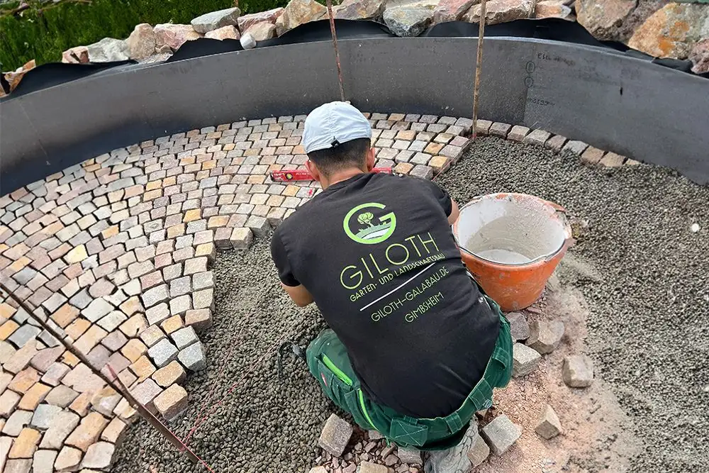 Handwerker im Giloth-Shirt verlegt buntes Natursteinpflaster in Bögen auf Splittbett für eine runde Pflasterfläche