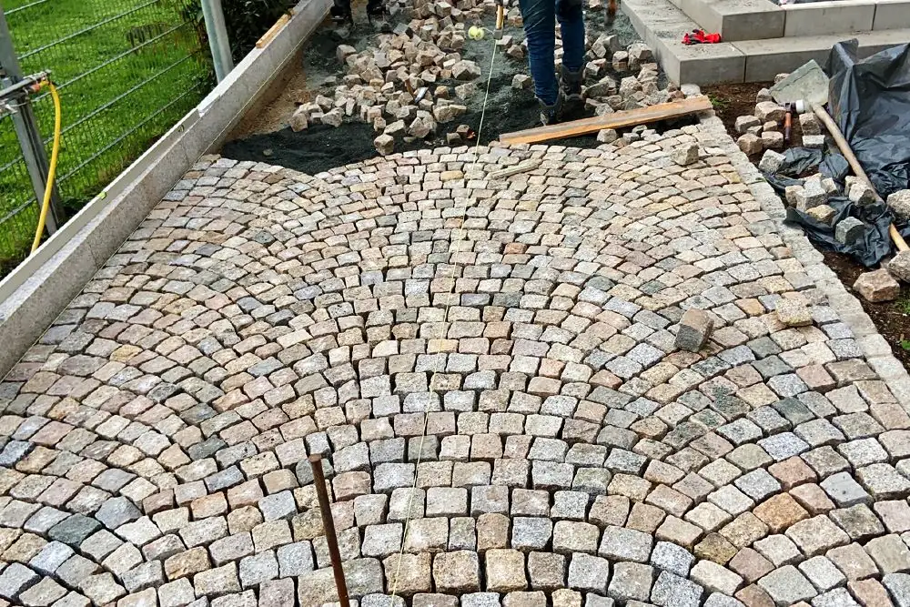 Buntes Natursteinpflaster wird im Segmentbogen verlegt und an der Schnur ausgerichtet, daneben liegen weitere Pflastersteine